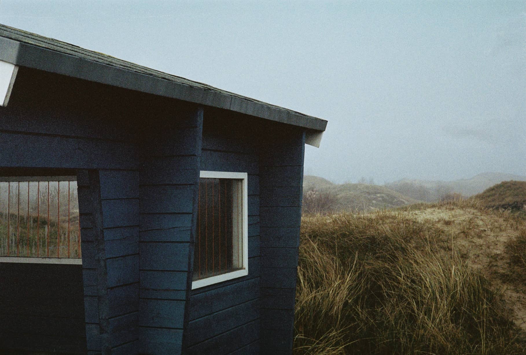 Dunkelblaue Holzhütte mit Fenstern vor Dünen und grauem Himmel